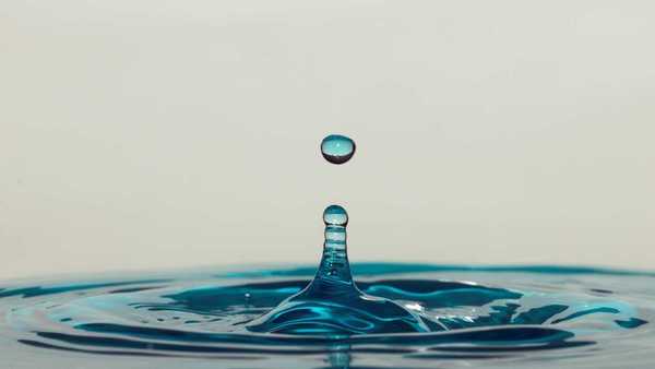 A close-up of a blue-tinted water droplet splashing into a surface, forming a ripple and upward splash against a neutral background.
