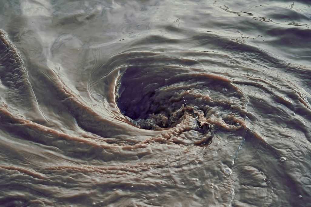 Close-up of murky water forming a swirling whirlpool, with brown and gray currents spiraling inward and rippled surface textures suggesting strong drainage or flow.